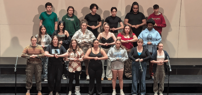 A choir of 19 singers standing in three rows on riser on a stage.