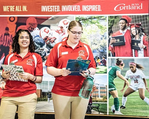 Two students in red polos with clipboards, standing in front of a SUNY Cortland banner while leading orientation