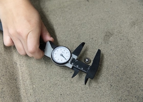 A student holds a pair of calipers and measures the diameter of a crater formed when a steel ball was dropped into the sand
