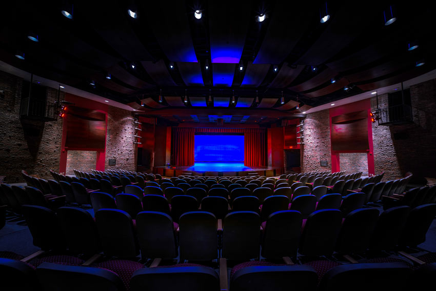 The Dowd Fine Arts mainstage theatre facing a large stage lit in blue, with red curtain. There are many auditorium seats in the foreground.